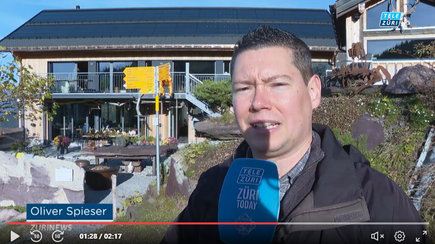 Reporter Oliver Spieser von TeleZüri steht mit Mikrofon vor dem Stockhus, einem modernen Eventlokal in den Schweizer Bergen, im Hintergrund das Gebäude und die umliegende Berglandschaft.