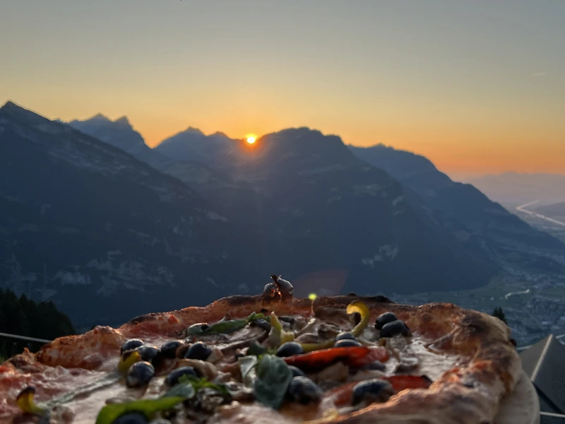 Eine knusprige Pizza mit frischen Zutaten im Vordergrund und ein spektakulärer Sonnenuntergang über den Glarner Alpen im Hintergrund.
