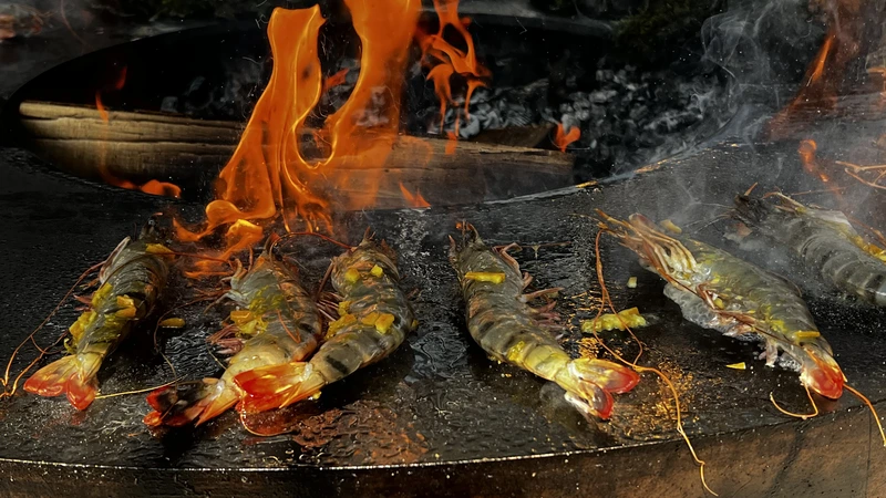 Saftige Riesengarnelen auf einer Grillplatte mit offenen Flammen im Hintergrund.