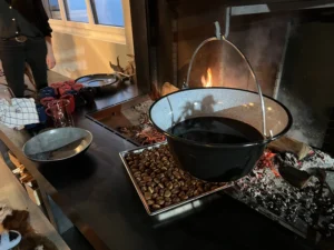Ein gemütlicher Kamin mit knisterndem Feuer, darüber hängt ein grosser Kessel. Im Vordergrund steht ein Tablett voller gerösteter Marroni auf einer Theke.