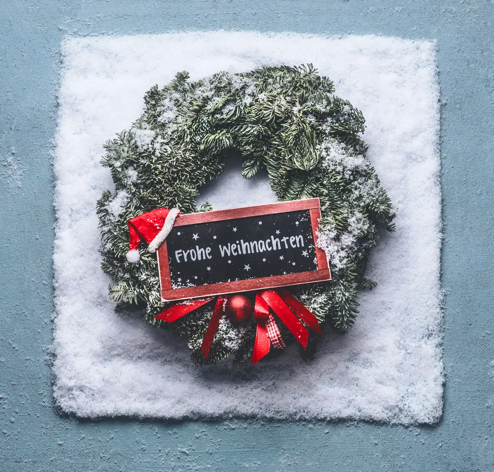 Weihnachtskranz mit roter Schleife und kleiner Tafel mit der Aufschrift „Frohe Weihnachten“, dekoriert mit Schnee – festliche Stimmung für den Weihnachtsgruss aus dem Stockhus auf der Fronalp.
